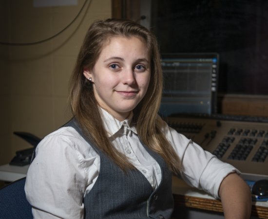 Crystal Hayner, with long brown hair, wearing a vest, sits by a computer set-up.
