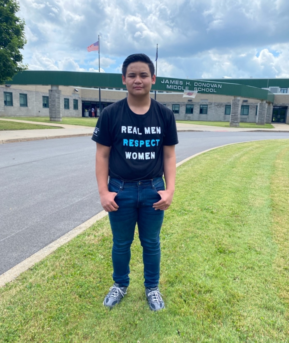 A young male student standing in front of Donovan Middle School wearing an AMEND Together shirt.