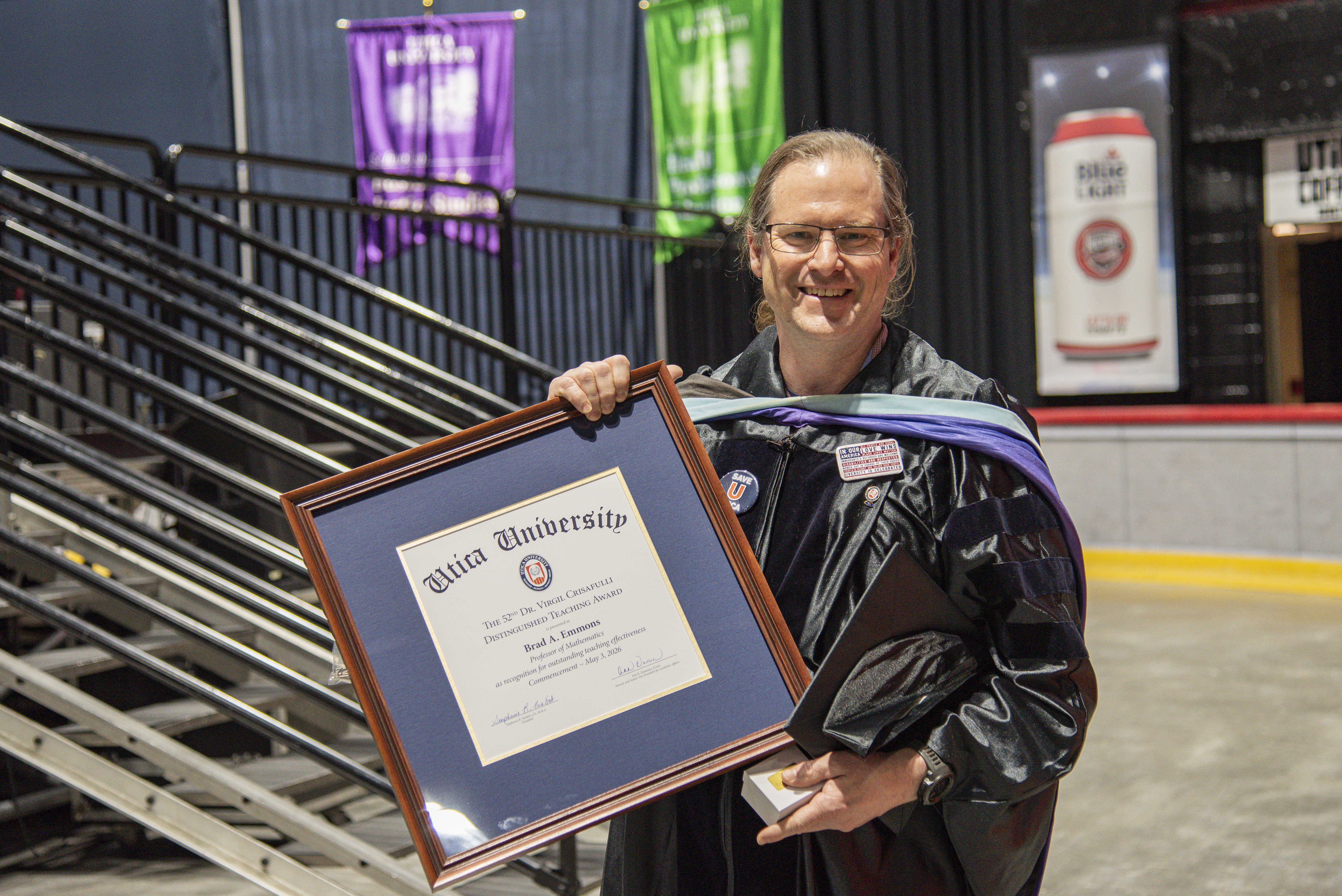Brad Emmons in commencement regalia, stands with the framed 2026 Crisafullli Award.