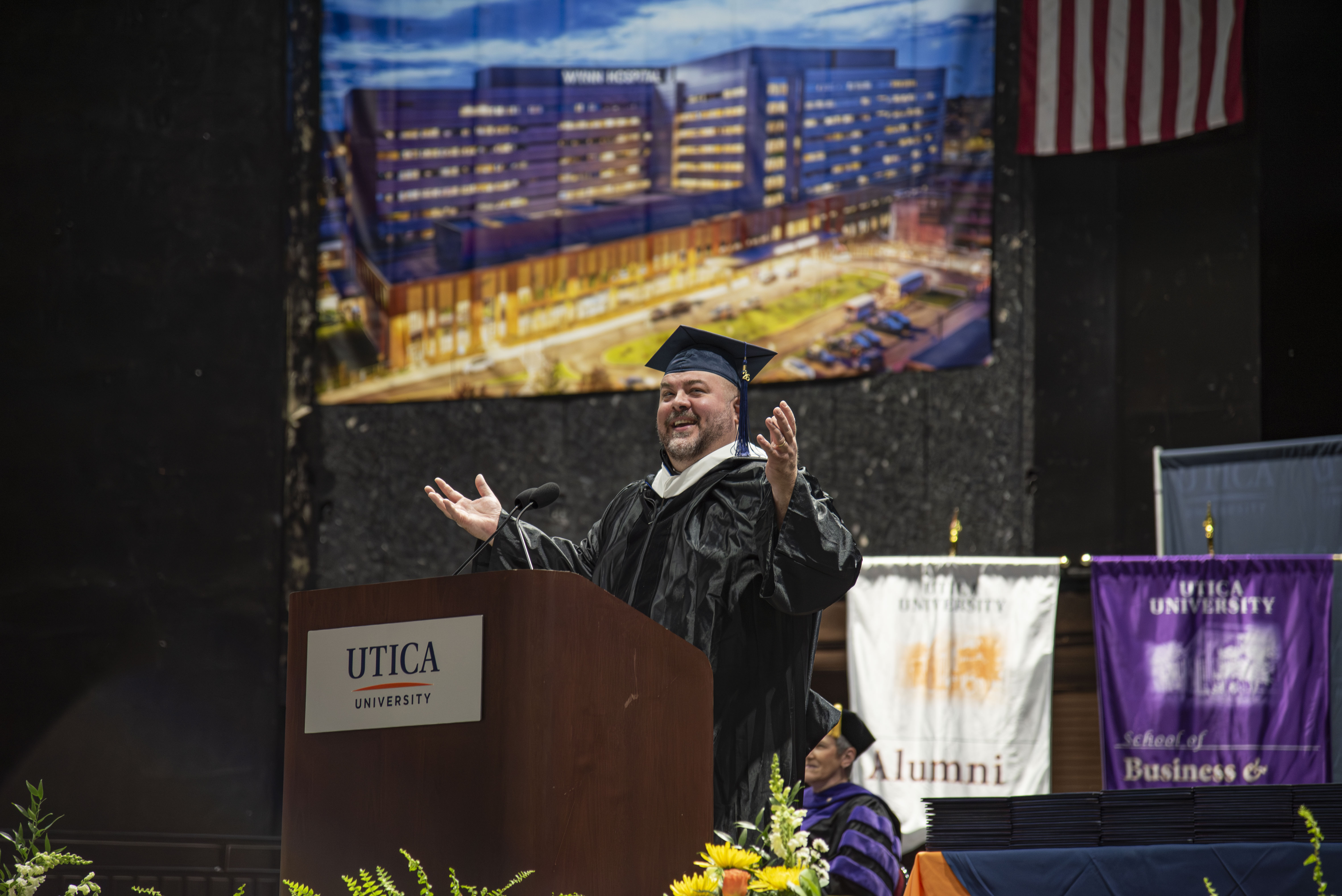 Harold Coleman G'17 in cap and gown, stands behind a lectern, with arms spread open, addressing the Graduate Class of 2026.