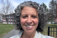 Donna Dolansky with shoulder length curly air stands on campus and smiles at the camera.