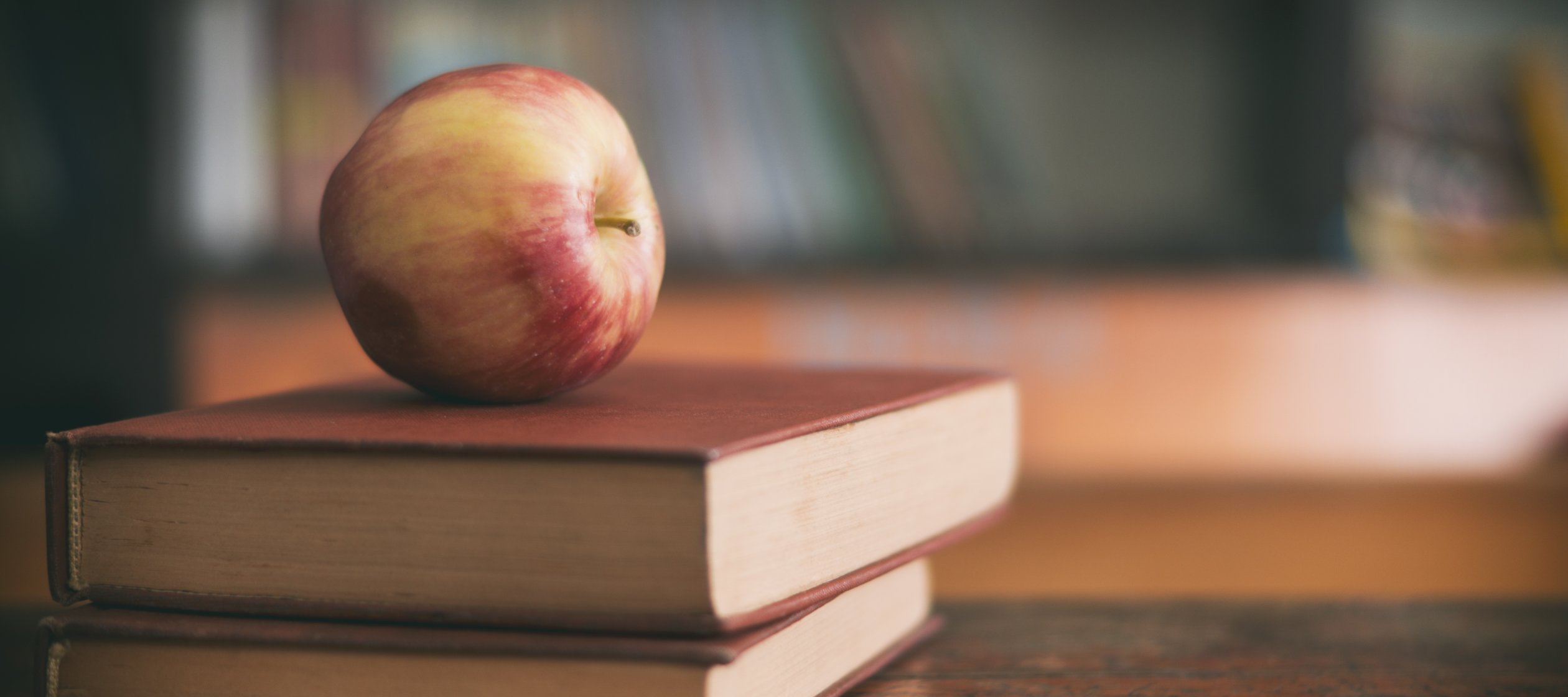Apple sitting on a pile of books on a classroom desk.