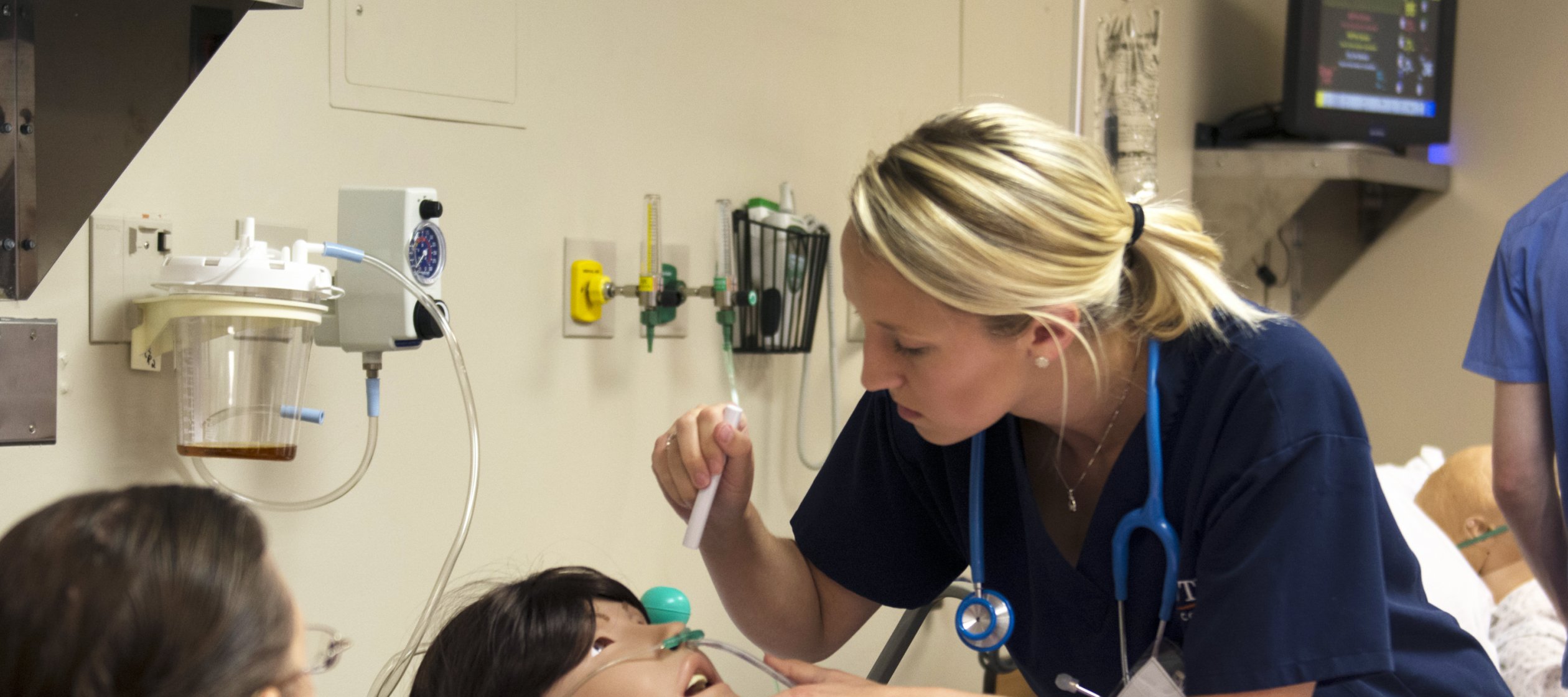 Nursing students in a lab simulation look over a mannequin in a hospital bed.