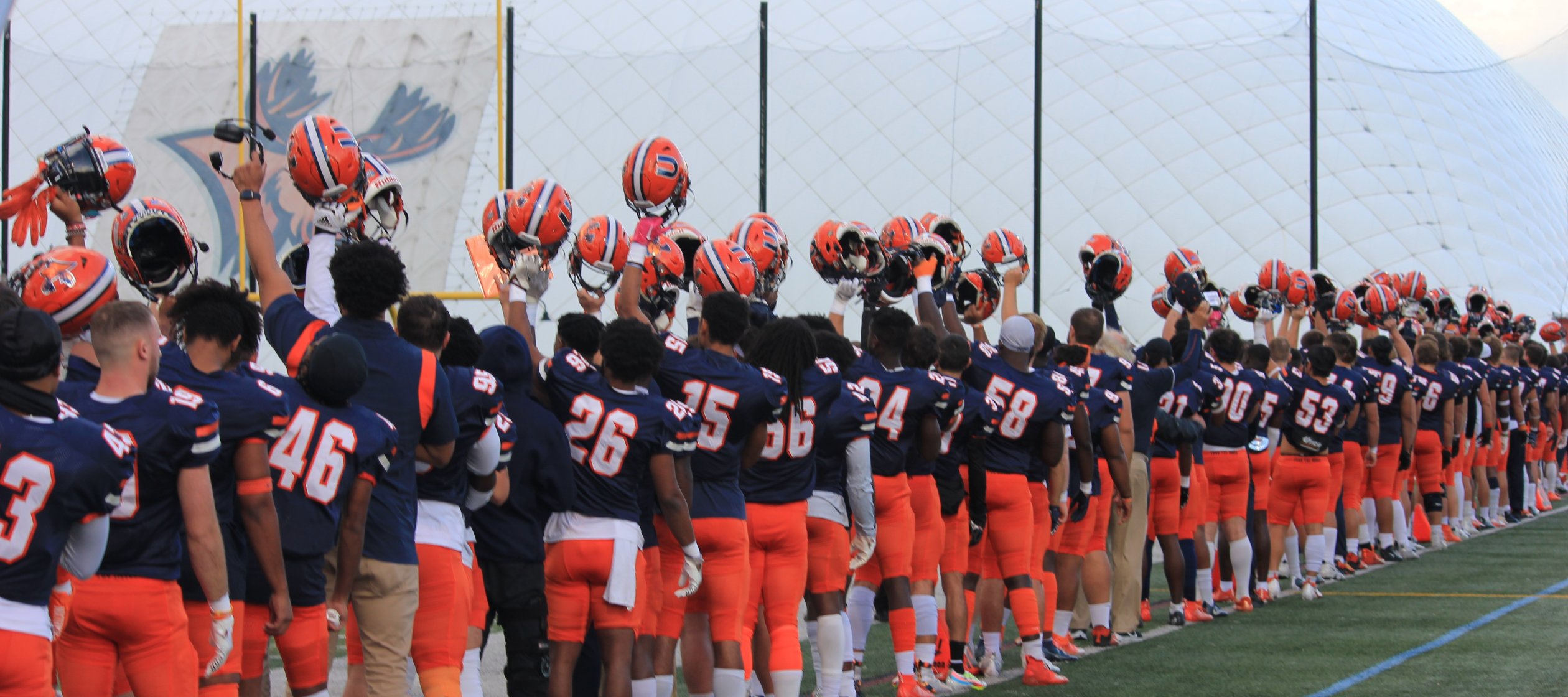 Members of the football team stand on the field outside the dome, holding their helmets up in the air.