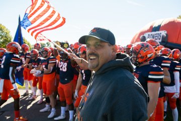 Coach Blaise Faggiano stands ahead of football players waving Pioneers flag