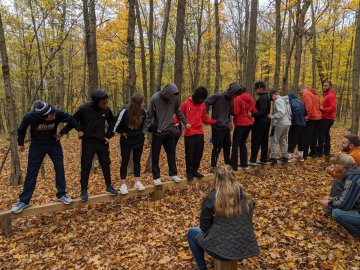 Students in the woods stand on a log holding hands.