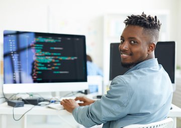 Cybersecurity student sits at computer, turning and smiling for camera.