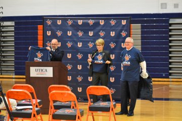 Dave Fontaine, at podium, holds up a Utica Pioneers branded t-shirt announcing three new varsity sports. President Casamento and Provost Pfannestiel stand nearby.