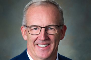 Greg Sankey, a man with short cut gray hair in glasses and blue suit, smiles at camera.