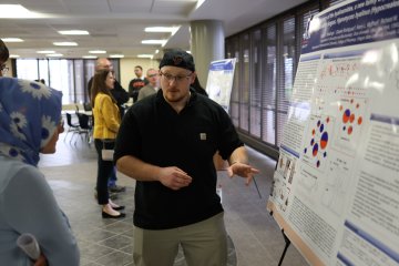 A student makes a presentation beside a poster at the 2024 Student Conference for Research, Professional Activities, and Creative Arts