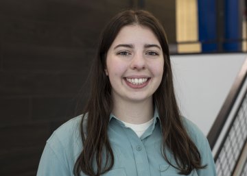 Sydney Kinsella, with long dark hair and wearing a blueish-green button-down top, looks at the camera and smiles.