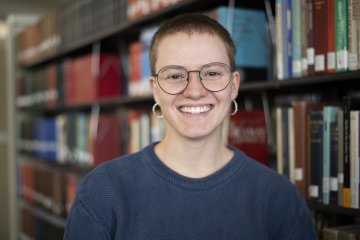 Glory Stevens in blue shirt and wearing glasses stands in front of library stacks.