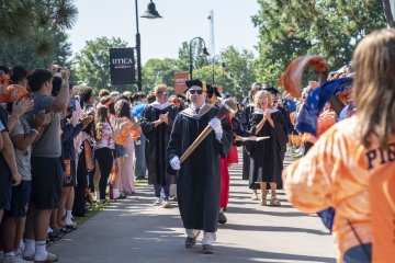 Professor Thomas Rossi acts as Macebearer, leading a procession of faculty down the sidewalk surrounded by cheering students.