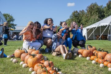 Students sit at the top of a big U made out of pumpkins on the grass of campus, making a U with their hands.