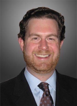 Man with dark hair and light brown beard in suit and tie smiles at camera.