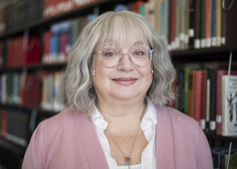 Janis Winn in glasses, white shirt and pink blazer stands in front of library stacks.