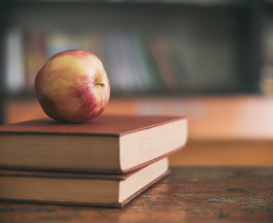 Apple sitting on a pile of books on a classroom desk.