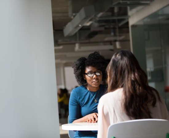 Woman at table speaking with another woman.