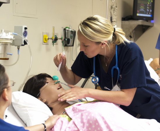 Nursing students in a lab simulation look over a mannequin in a hospital bed.