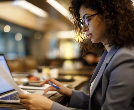 Woman in suit jacket and glasses holds a sheet of paper in one hand while typing on a laptop with the other hand.