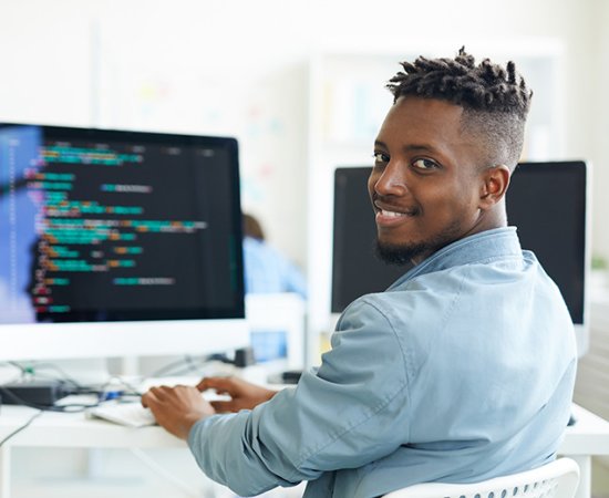 Cybersecurity student sits at computer, turning and smiling for camera.