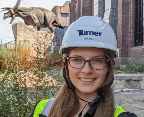 Nicole Zalewski, wearing glasses and a construction helmet, stands in front of an old brick building with a statue of a triceratops out front.