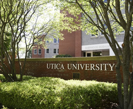 The words Utica University against a brick wall in front of a residence hall.