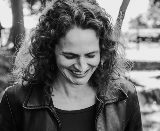 Black and White image of Jessica Cuello in leather jacket, smiling, walking through woodded area.