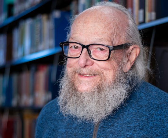 John Cormican, with gray beard and hair down to his shoulders, wearing black glasses and a blue sweater, stands in front of book shelves and smiles.