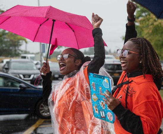 Two students under umbrellas shout excitedly with arms in the air and smiles on their faces as they greet new students.