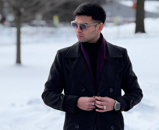 Muhammad Hamza Bilal in sunglasses, buttons up black coat while walking against a snowy backdrop.