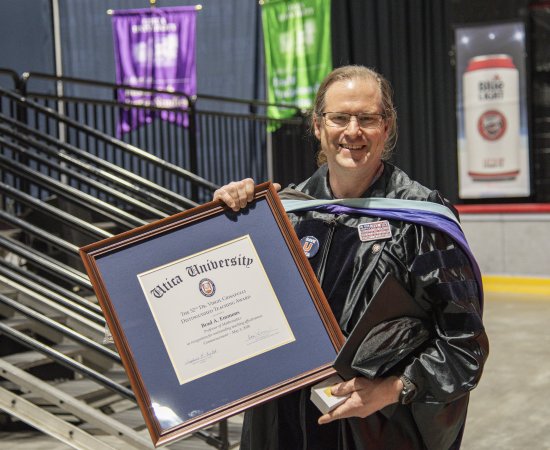 Brad Emmons in commencement regalia, stands with the framed 2026 Crisafullli Award.