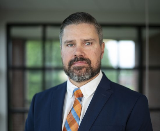 Matthew Marmet, with beard, blue shirt and orange and blue tie, stands in front of glass windows.