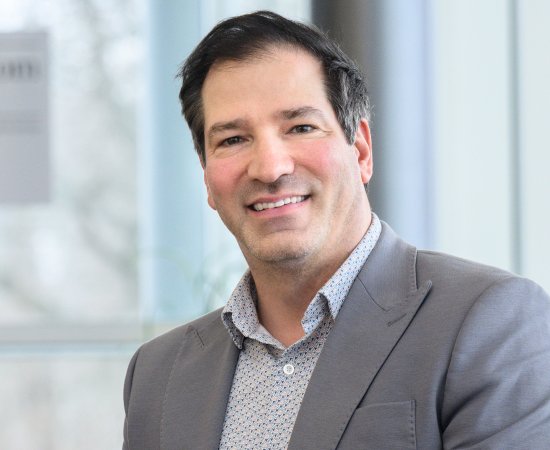 Dr. Christopher Riddle in gray suit jacket sits in front of a wall of windows and smiles.