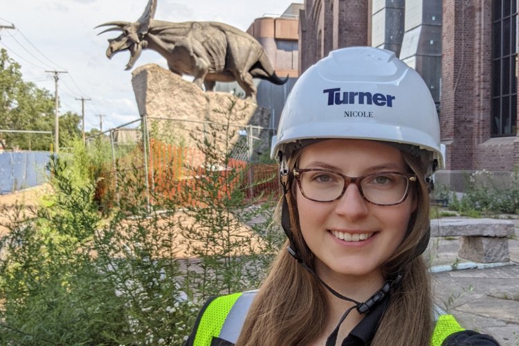 Nicole Zalewski, wearing glasses and a construction helmet, stands in front of an old brick building with a statue of a triceratops out front.