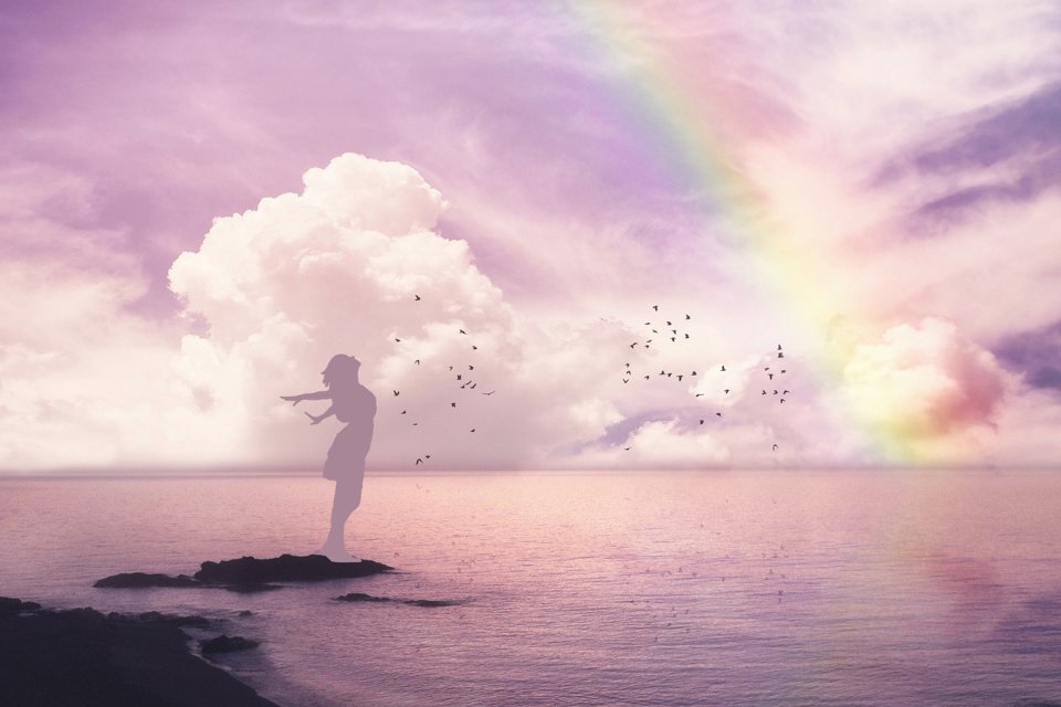 Silhouette standing on rock amid water, at peace as a rainbow shines in the sky.