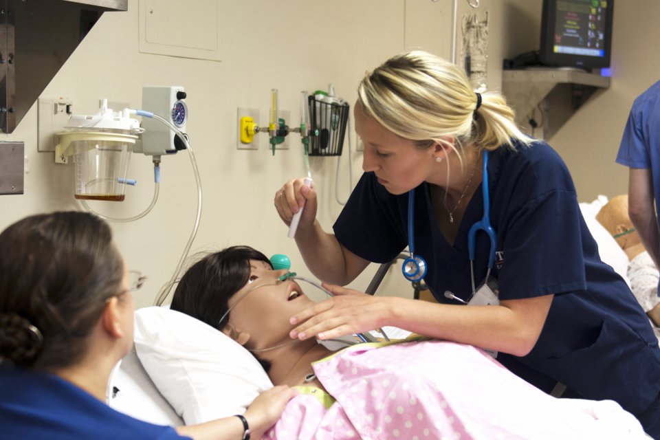 Nursing students in a lab simulation look over a mannequin in a hospital bed.