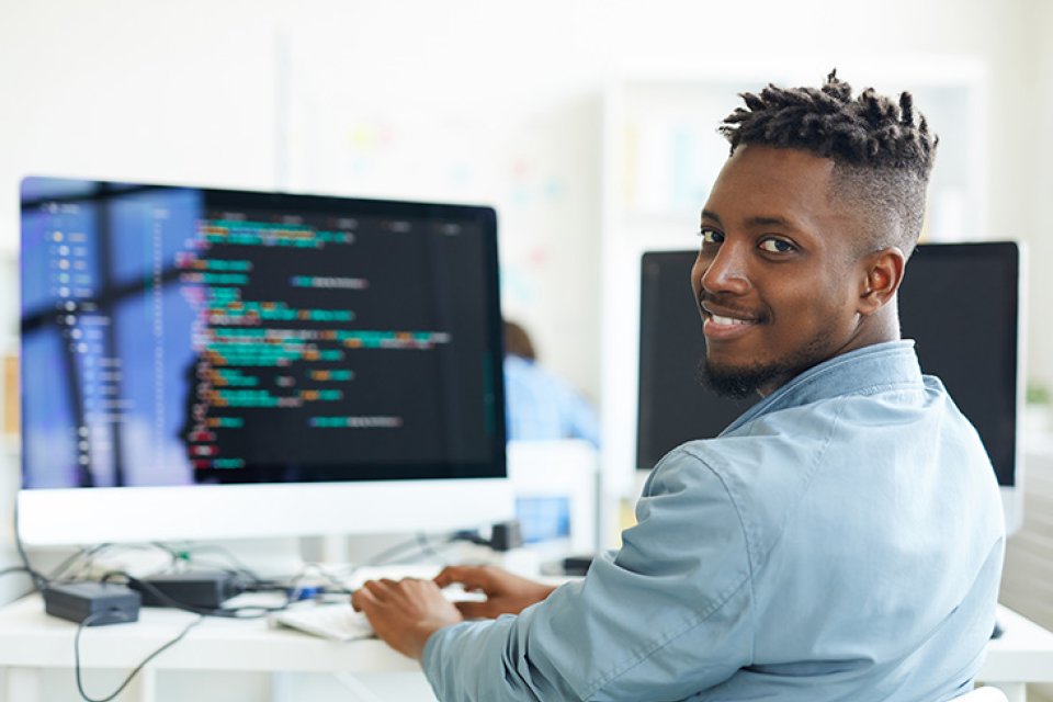 Cybersecurity student sits at computer, turning and smiling for camera.