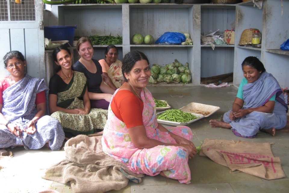 A group of Indian women in saris sit on the floor of a kitchen, smiling at the camera. Behind them are shelves of produce, as well as plates of beans.