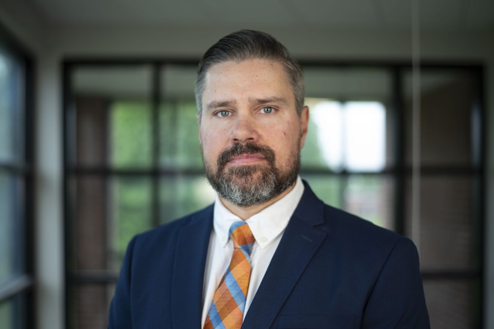 Matthew Marmet, with beard, blue shirt and orange and blue tie, stands in front of glass windows.