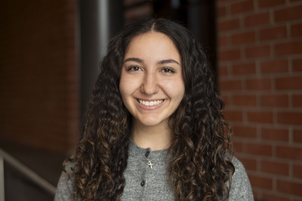 Olivia D’Ambro, with long, dark, curly hair and a gray shirt, stands in front of a brick wall and smiles.