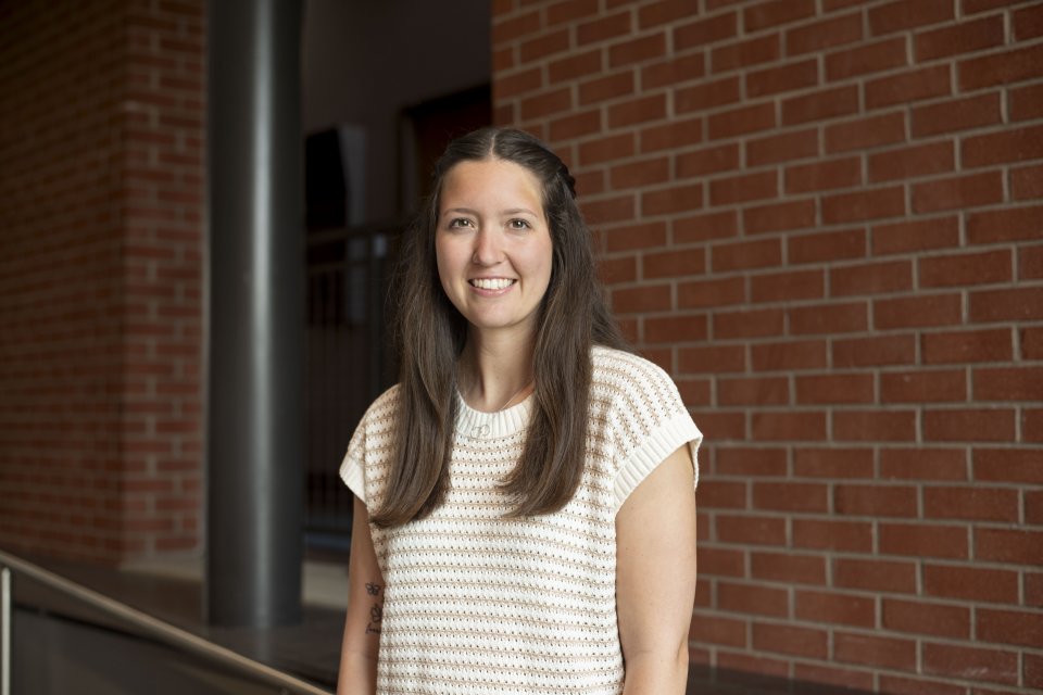 Kyla Reed, with long brown hair past her shoulders, wearing striped short sleeve shirt, stands in front of a brick wall and smiles.